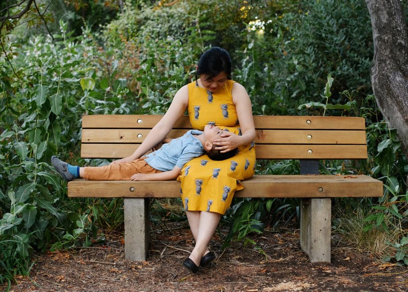 mom sitting on a bench with young son laying in her lap during summer family photos in davis ca