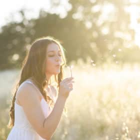 senior in high school blowing a dandelion in a field during golden hour