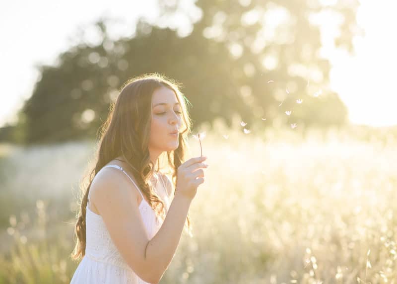 senior in high school blowing a dandelion in a field during golden hour