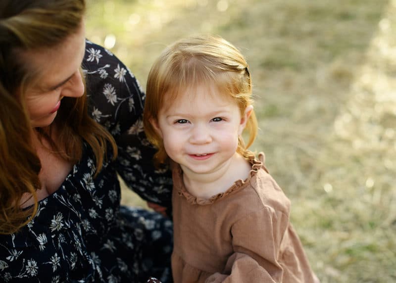 young girl sitting on pregnant mom's lap and looking up at the photographer smiling