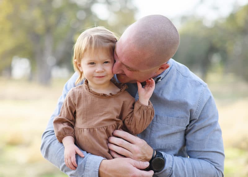 young girl looking at the photographer while dad kisses her cheek during maternity photos in folsom california