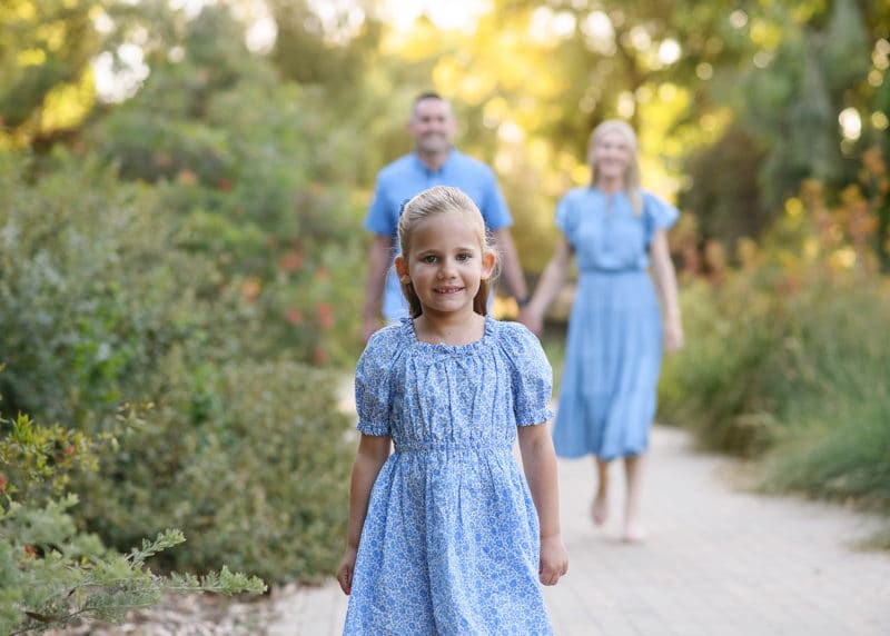 young girl walking toward the photographer with mom and dad holding hands behind them