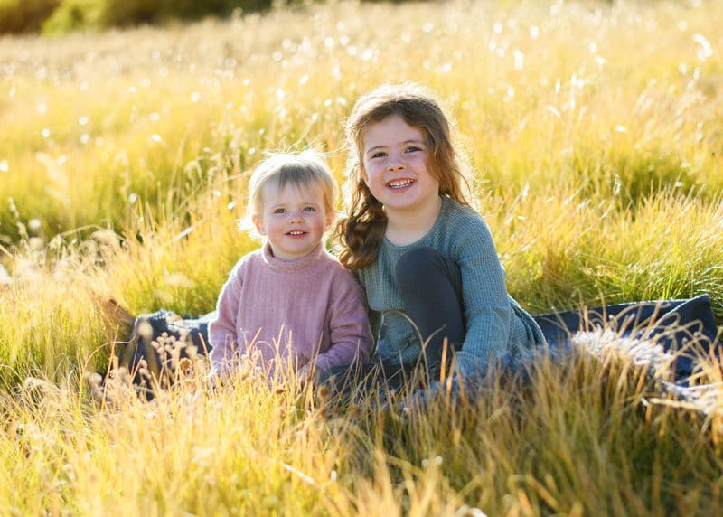 two young sisters sitting in a field by the mountains of lake tahoe looking at the photographer and smiling