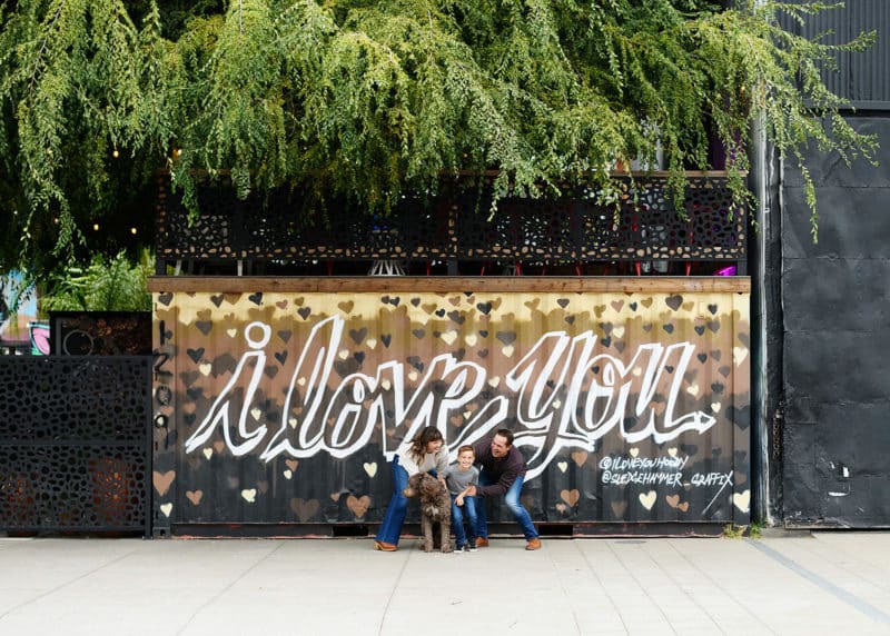 family of three standing in front of a mural that says "i love you" with family dog in downtown sacramento california