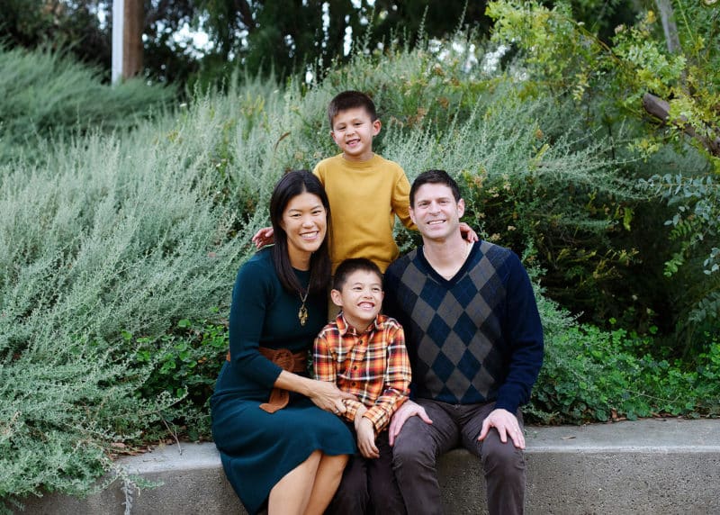 family of four wearing fall colors and sitting on a concrete bench in front of foliage during fall family photos in davis california