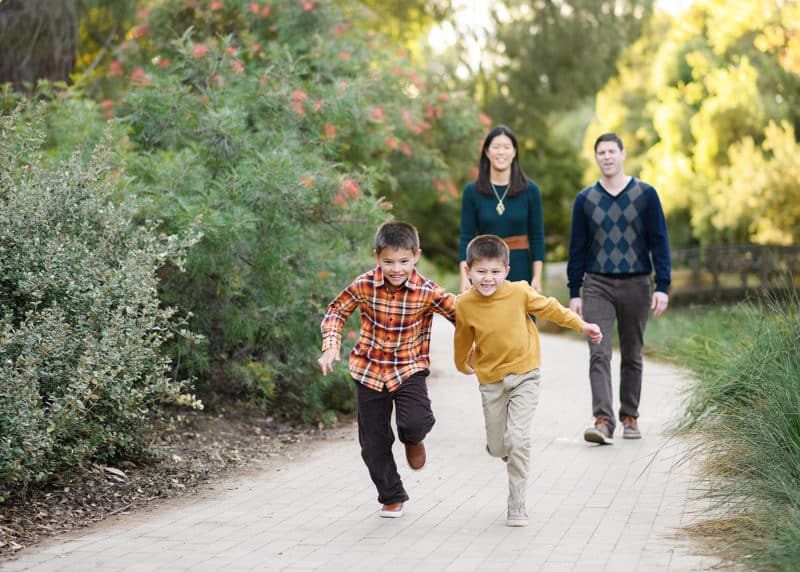 two young boys running on a path while mom and dad watch in the background during fall family photos with davis photographer jill carmel