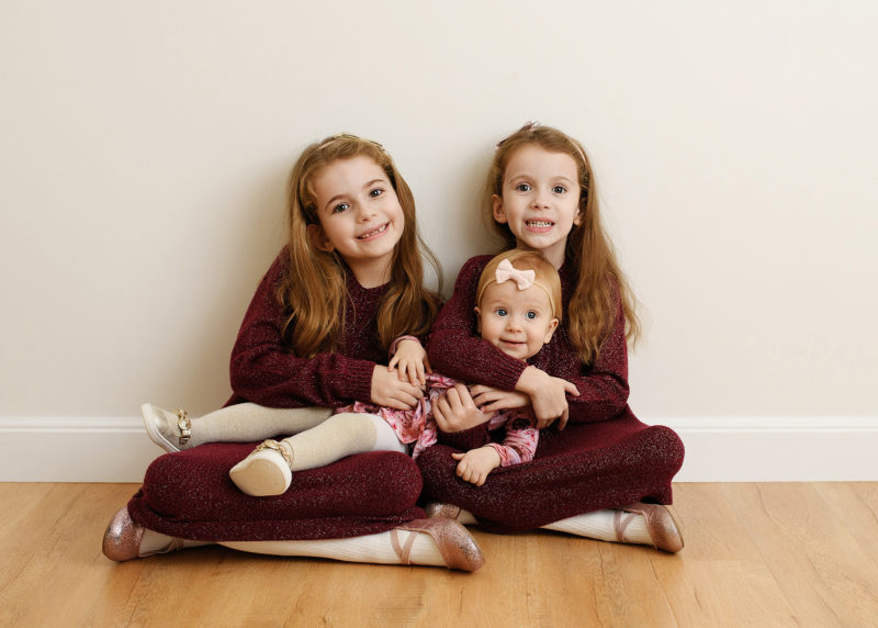 three young sisters sitting against a wall and smiling during family studio session with sacramento photographer jill carmel